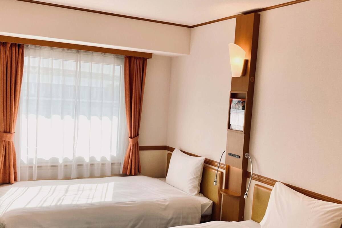 A hotel room with two beds, a large window with sheer curtains, a bedside table with a lamp, and a wall-mounted shelf.