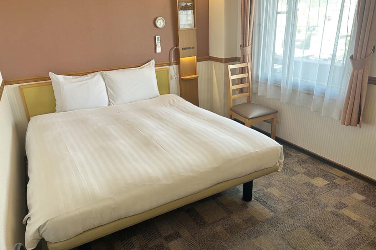 Quarto de hotel com uma cama grande, uma cadeira de madeira, uma mesa de cabeceira e uma janela com cortinas.