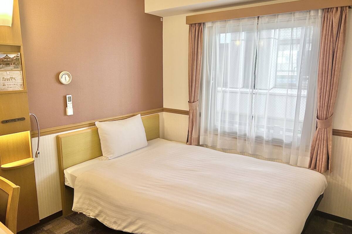 Quarto de hotel com cama, mesa de cabeceira, aquecedor fixado na parede e janela com cortinas leves.
