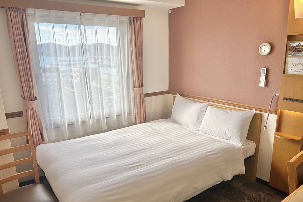 Quarto de hotel com cama, cadeira, janela com cortinas, mesa de cabeceira e relógio fixado na parede.
