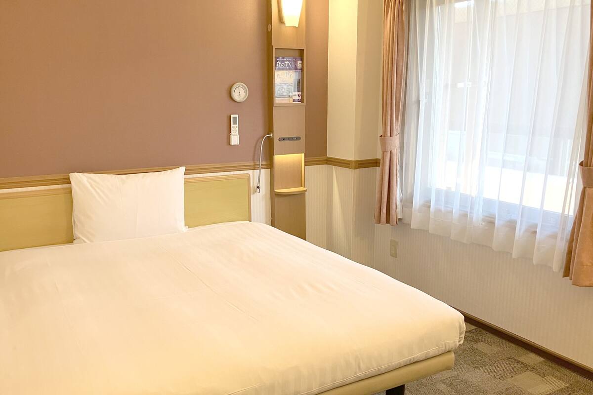 Quarto de hotel com uma cama grande, mesa de cabeceira, relógio e janela com cortinas.