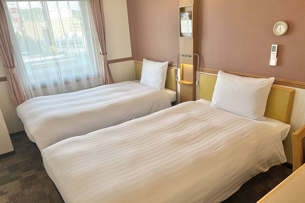 Quarto de hotel com duas camas de solteiro, uma janela com cortinas, um termostato e uma mesa de cabeceira.