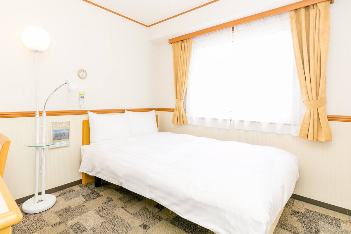A hotel room with a bed, a desk with a chair, a lamp, and a window with curtains.