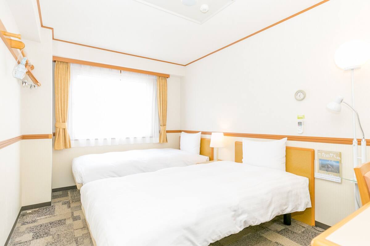 A hotel room with two beds, a desk, a chair, a lamp, a clock, and a window with curtains.