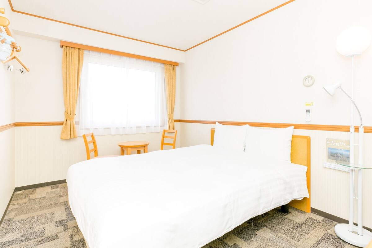 A hotel room with a bed, a desk, a chair, a lamp, and a window with curtains.