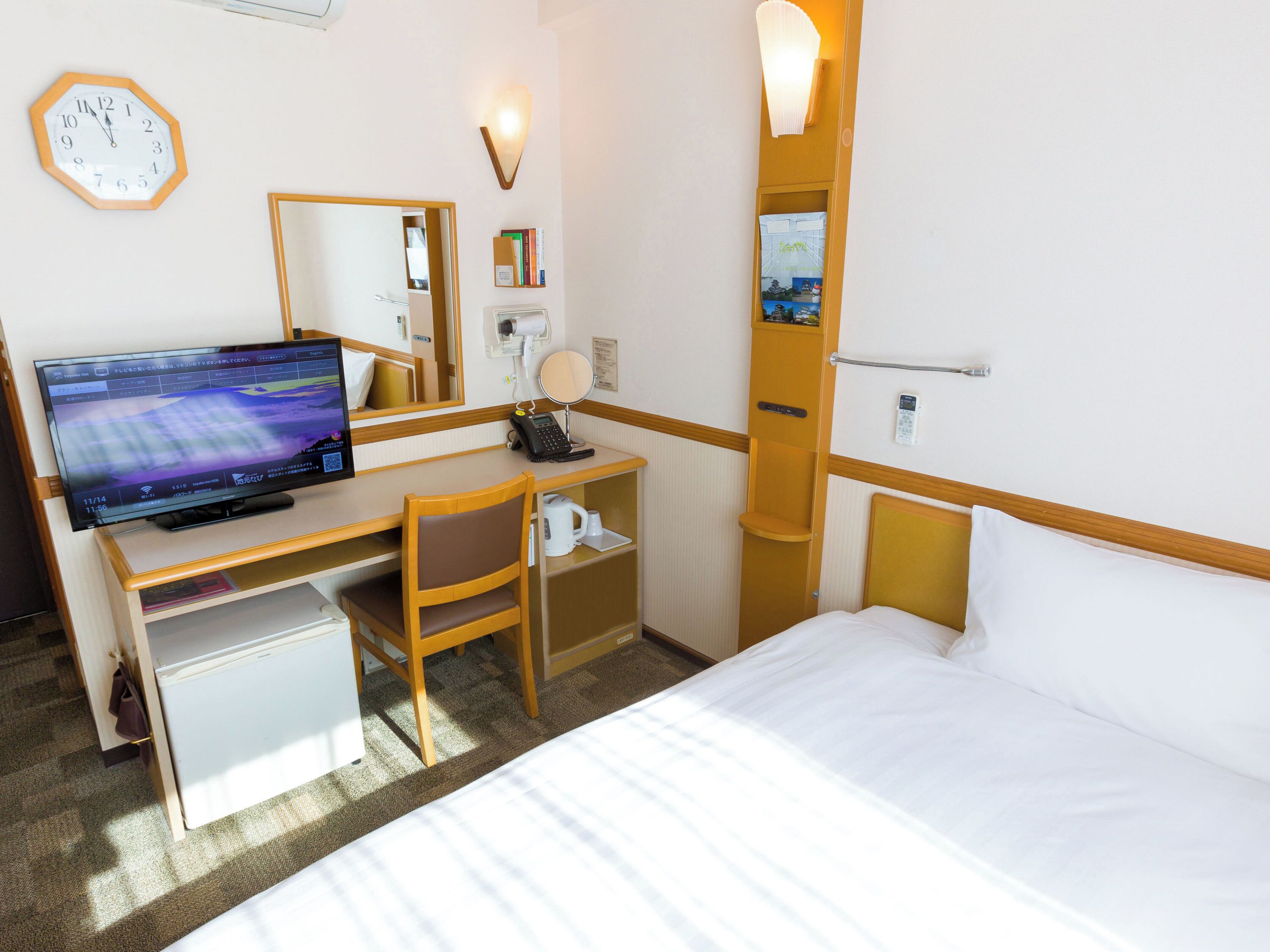 Habitación de hotel con cama, escritorio con computadora, silla, refrigerador pequeño, mesita de noche, lámpara y un reloj en la pared.