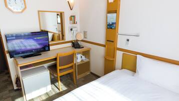 A hotel room with a bed, a desk with a computer, a chair, a small refrigerator, a bedside table, a lamp, and a clock on the wall.