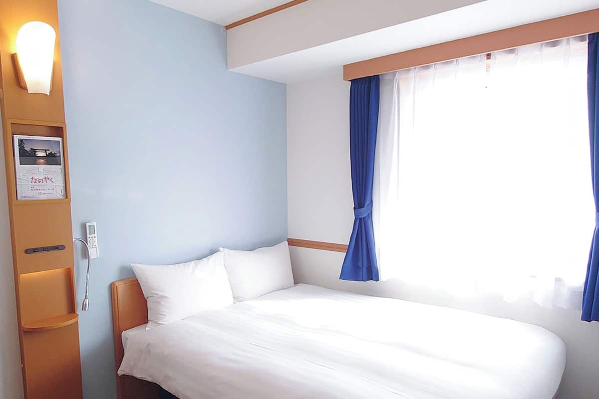 A hotel room with a bed, a bedside table, a lamp, a wall-mounted TV, and a window with curtains.