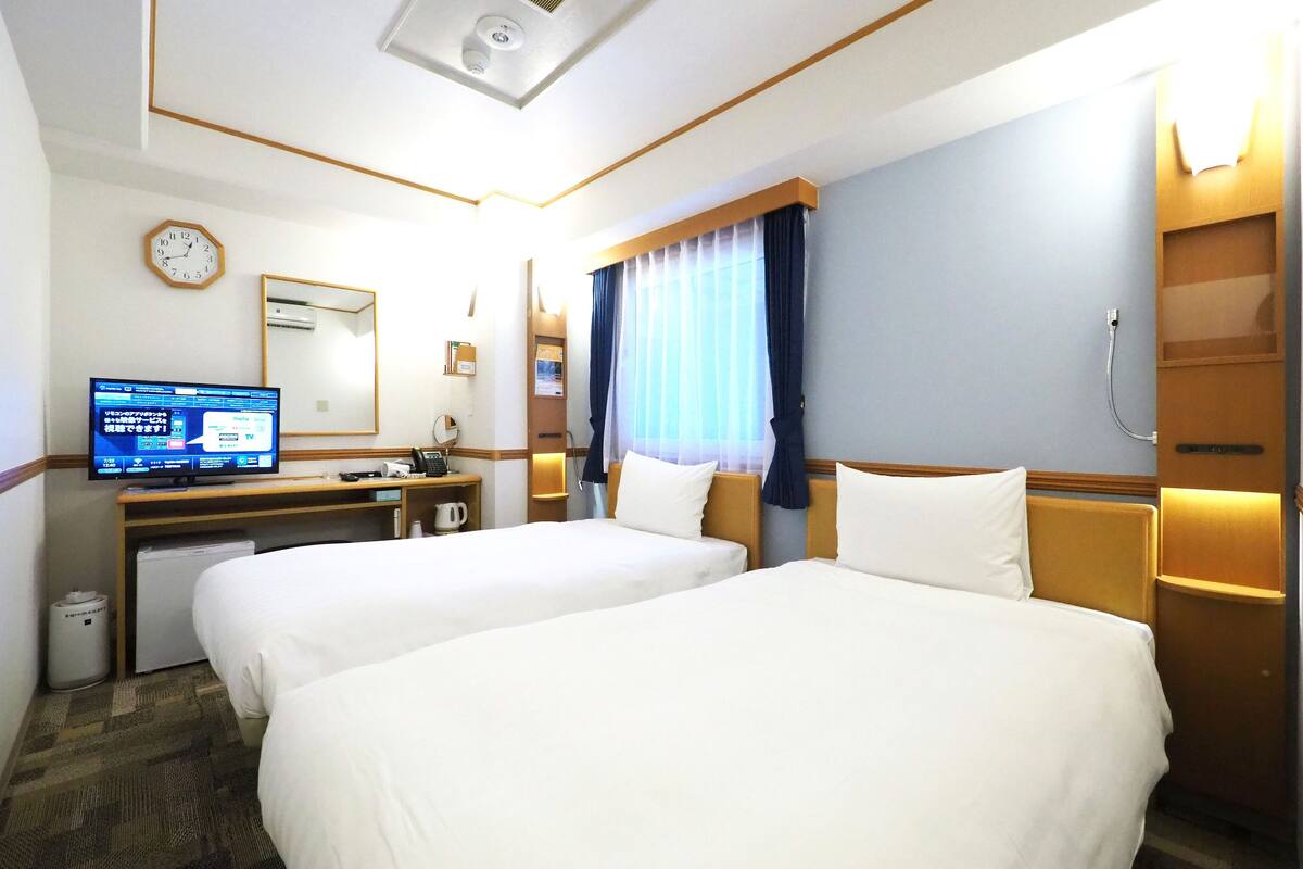 A hotel room with two beds, a desk with a computer, a clock, and a window with curtains.