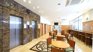 Un hall d’hôtel aux murs de marbre, avec des ascenseurs, une horloge, un distributeur automatique et un espace détente comprenant des tables et des chaises rondes.