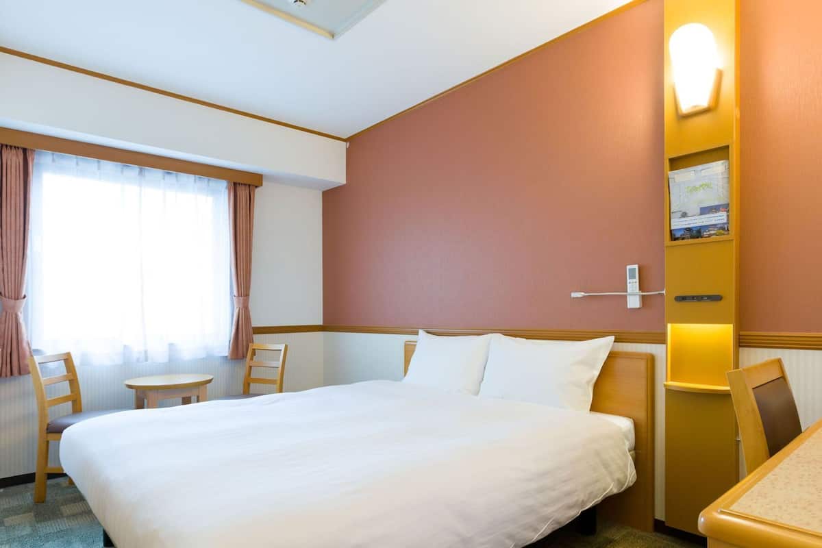 A hotel room with a large bed, a desk, two chairs, a small table, a wall-mounted phone, and a window with curtains.