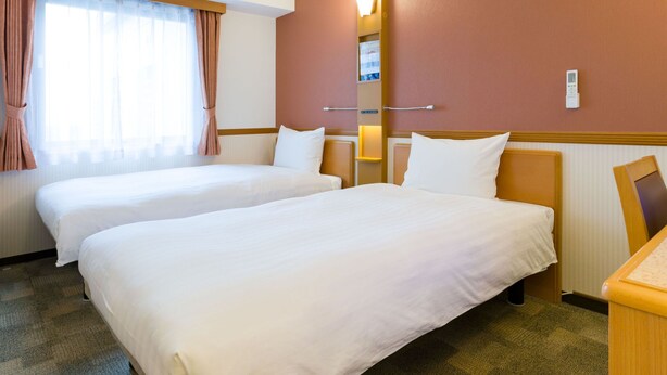 Ein Hotelzimmer mit zwei Betten, einem großen Fenster mit Vorhängen, einer Wandlampe und einem kleinen Schreibtisch.
