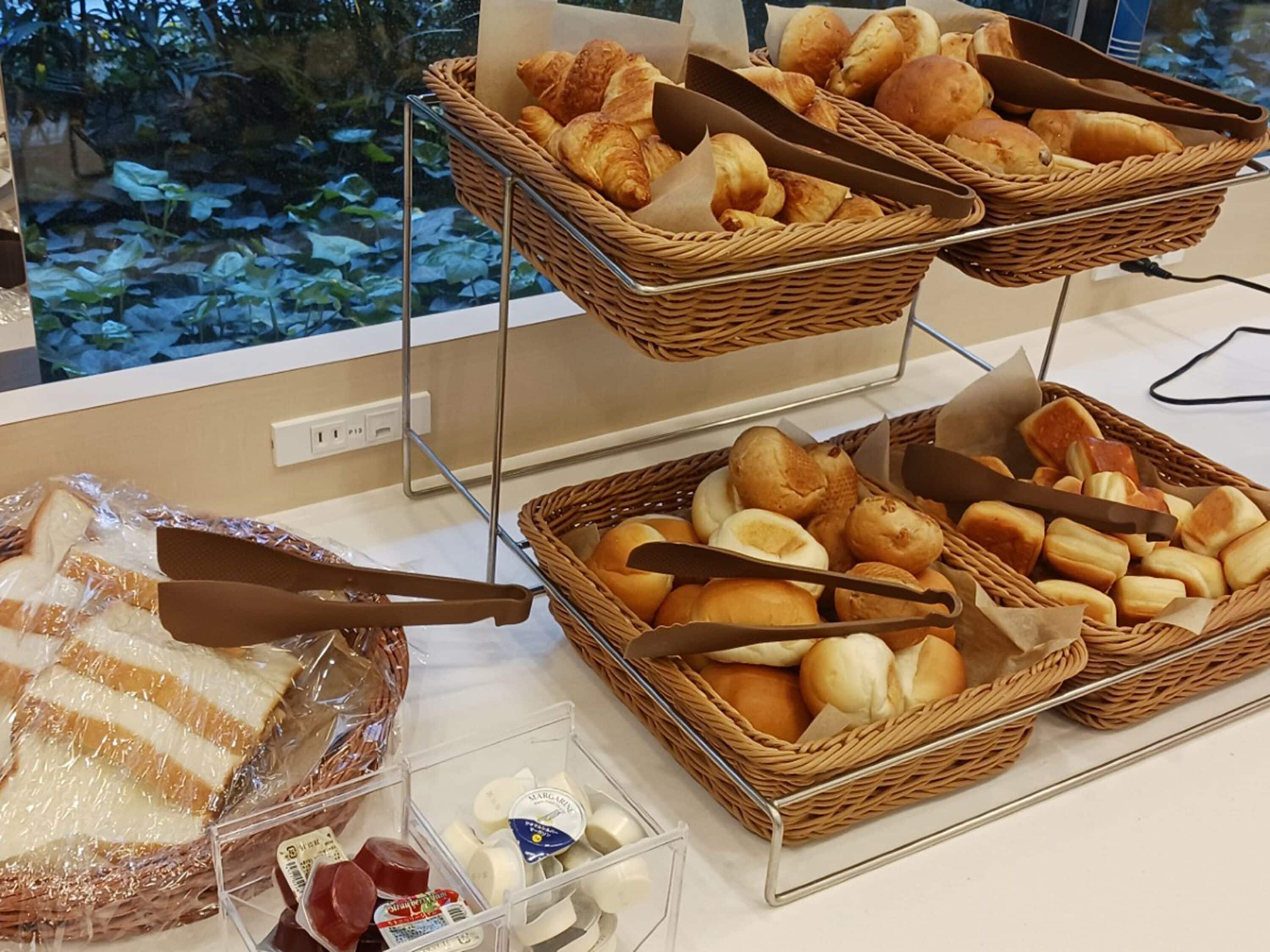 Taula de buffet amb diferents pa i pastissos en cistos de vímet, una barra de pa embolcallada en plàstic, i alguns aliments envasellats.