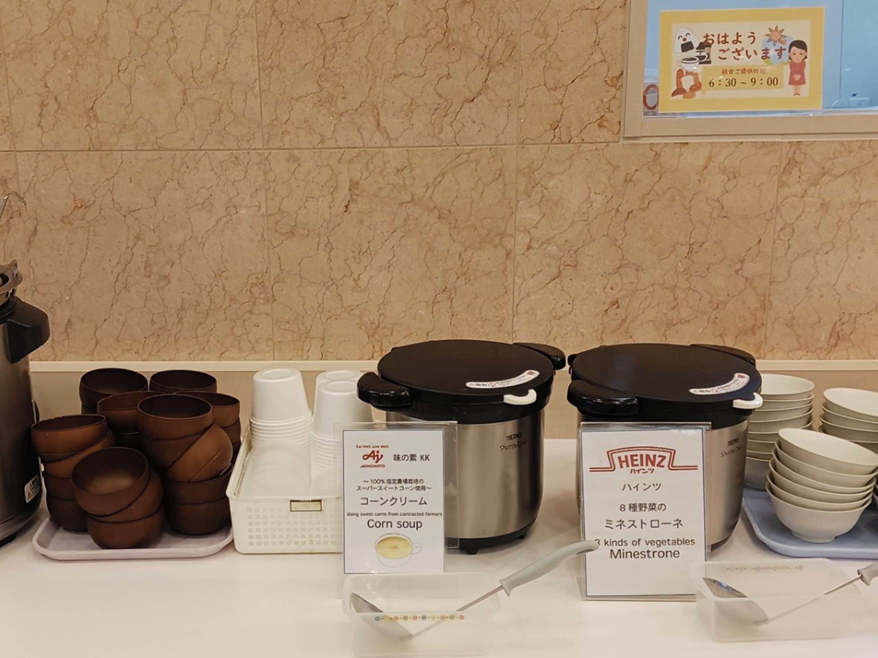 A buffet area with various food items and containers, including a sign for corn soup and a sign for minestrone.
