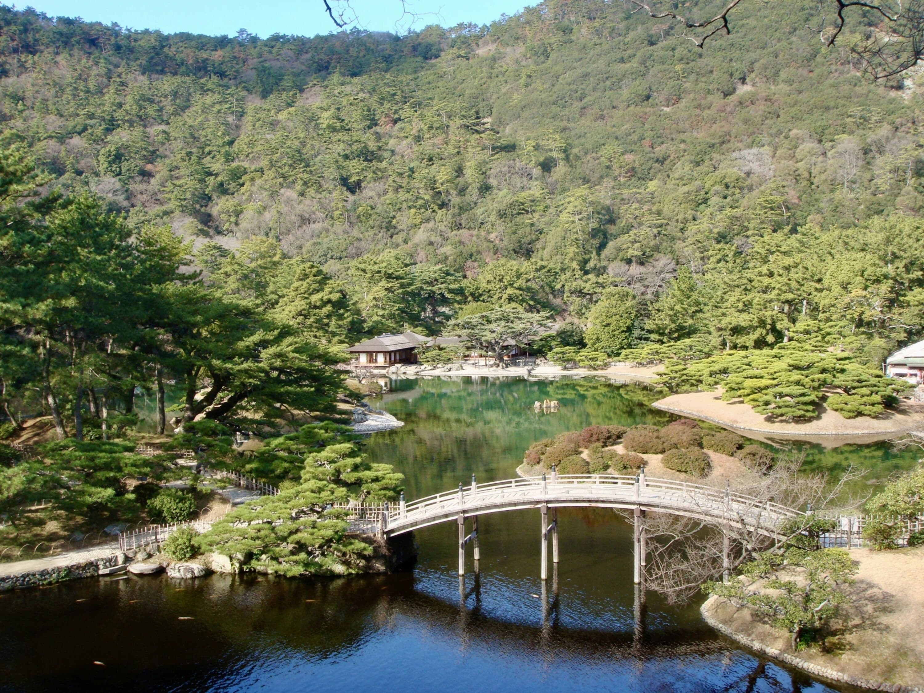Un jardín japonés tradicional con un puente de madera sobre un estanque, rodeado de abundante vegetación y un telón de fondo montañoso.