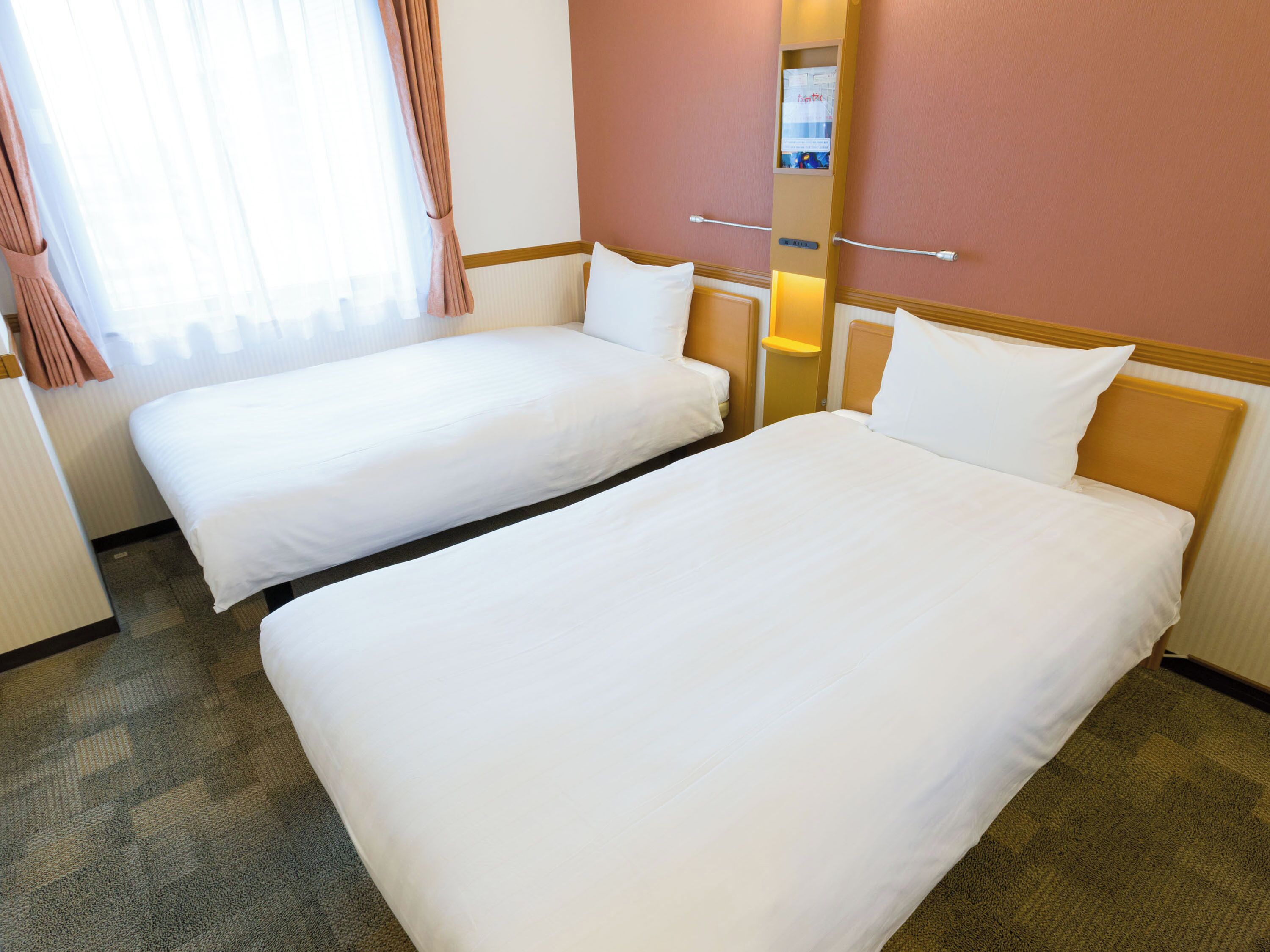 Una habitación de hotel con dos camas individuales, una mesita de noche pequeña, cortinas en una ventana y una lámpara de pared.