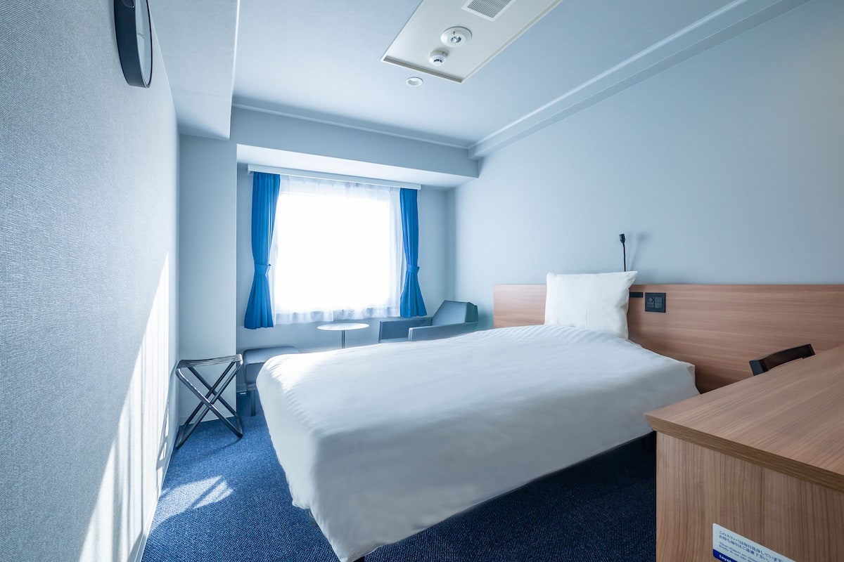 Habitación de hotel con una cama grande, un escritorio, una silla, una mesita y una ventana con cortinas.