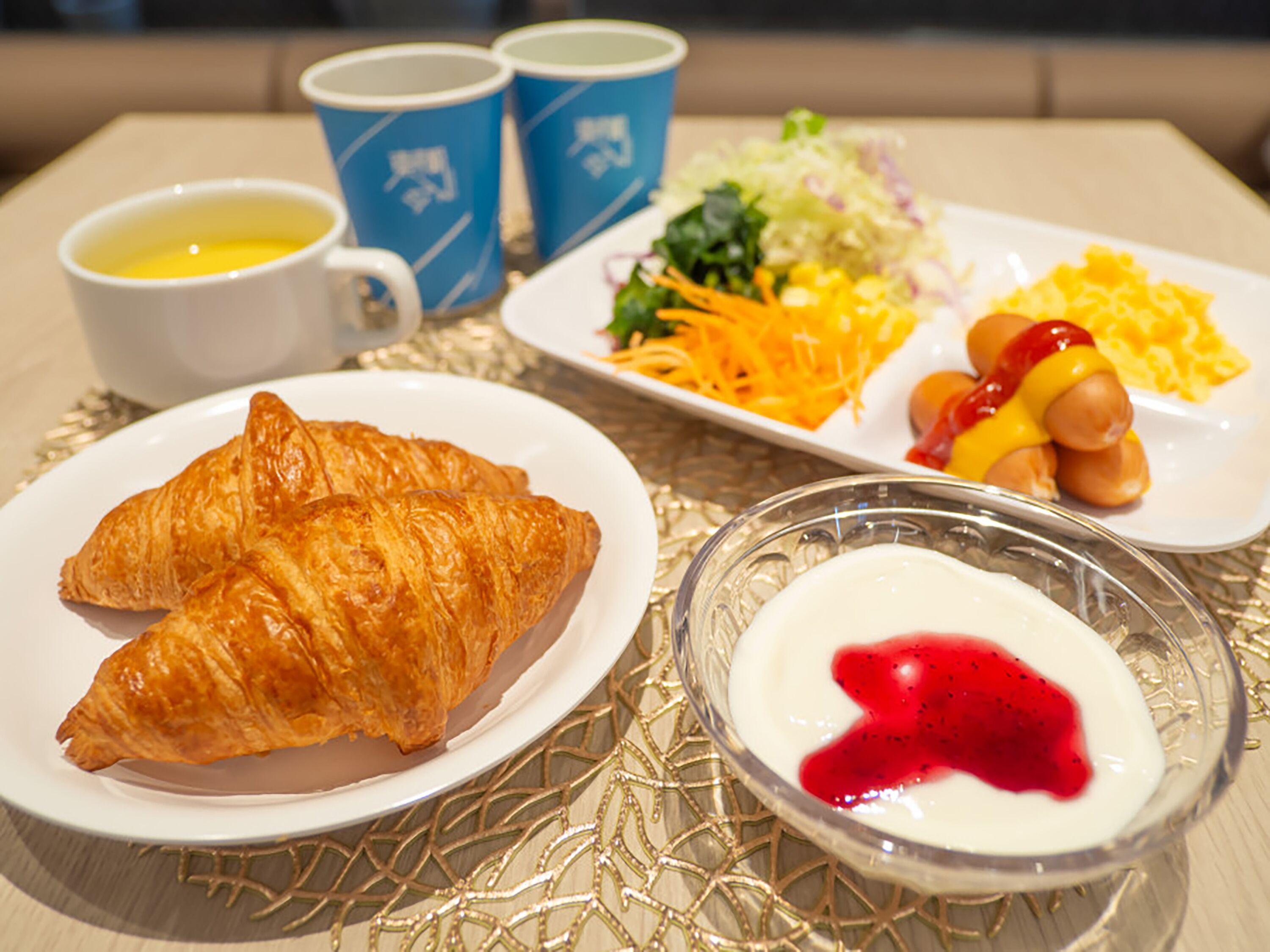 A breakfast setting with croissants, a salad, sausages, and a bowl of yogurt with red jam.