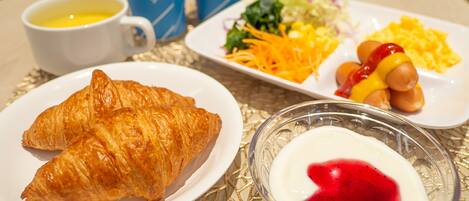 A breakfast setting with croissants, a salad, sausages, and a bowl of yogurt with red jam.