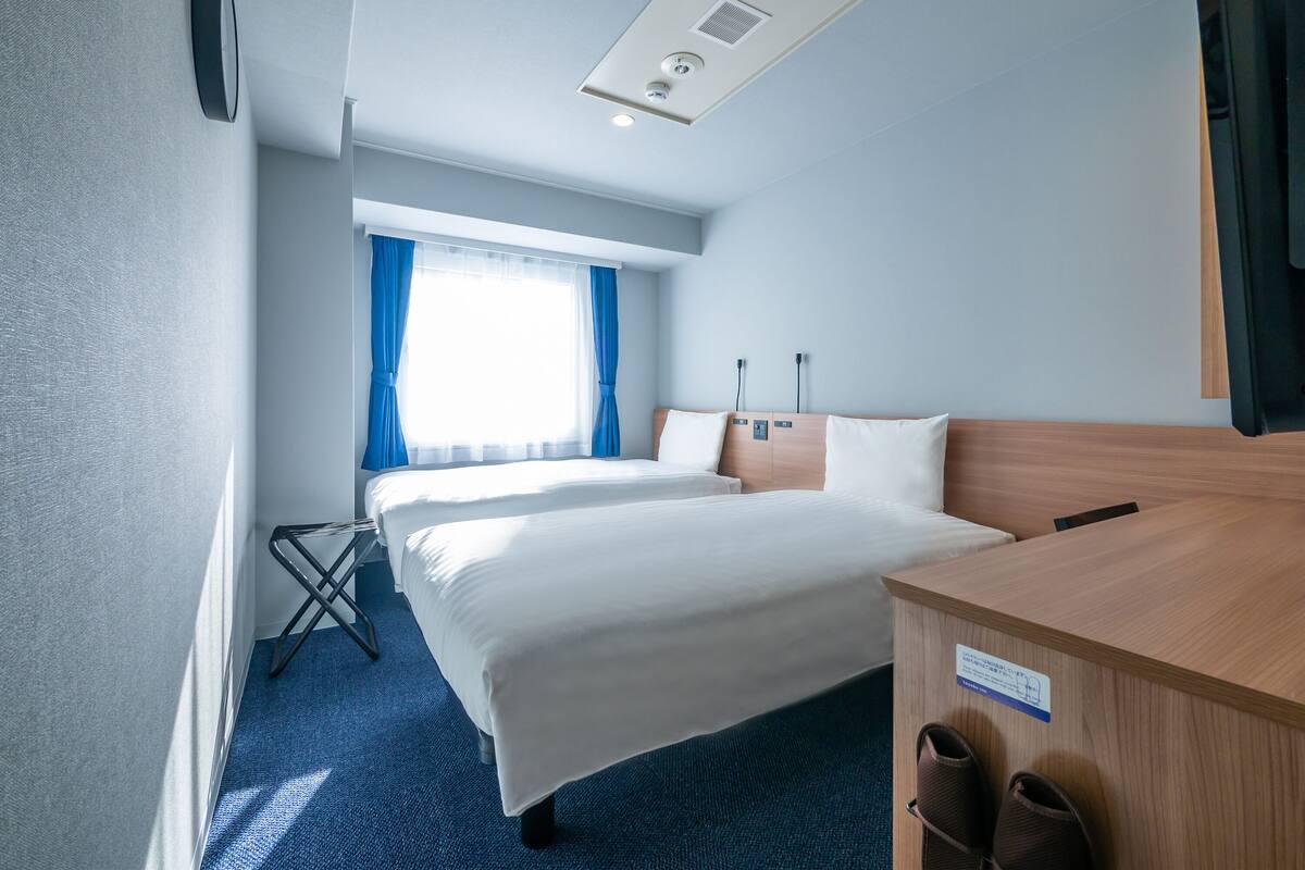Habitación de hotel con una cama grande, un escritorio y una silla. Hay una ventana con cortinas, una lámpara de techo y un televisor montado en la pared.