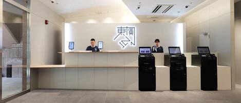 A modern reception desk with two staff members, a digital signboard, and a self-service kiosk.