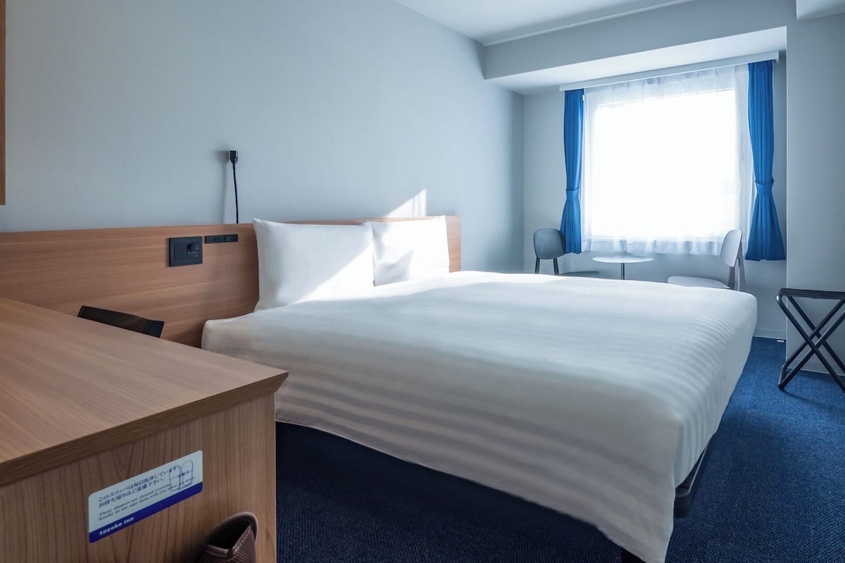 Habitación de hotel con una cama grande, un escritorio de madera, una silla, una mesita y una ventana con cortinas.