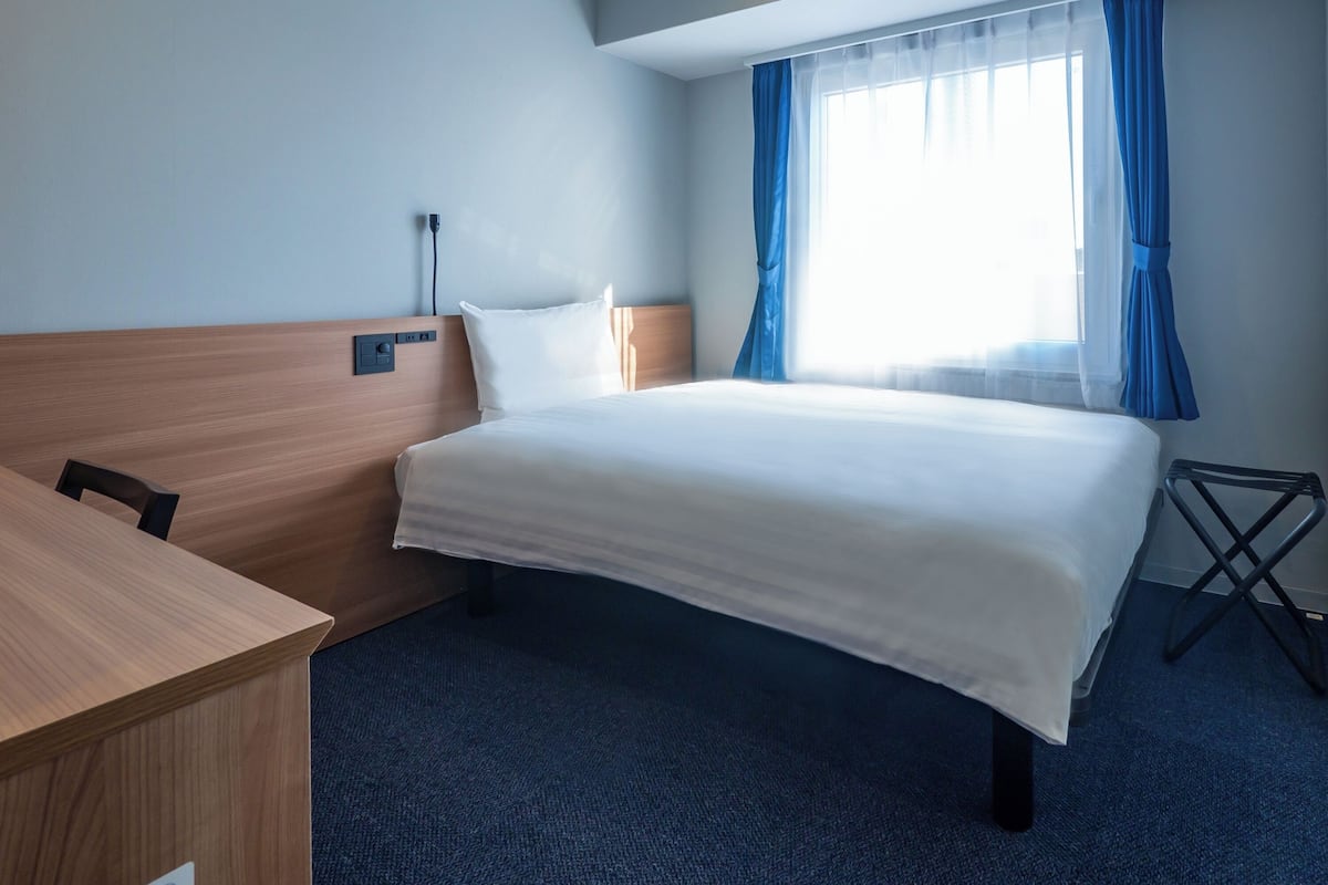 Habitación de hotel con cama, escritorio, silla, lámpara y ventana con cortinas.