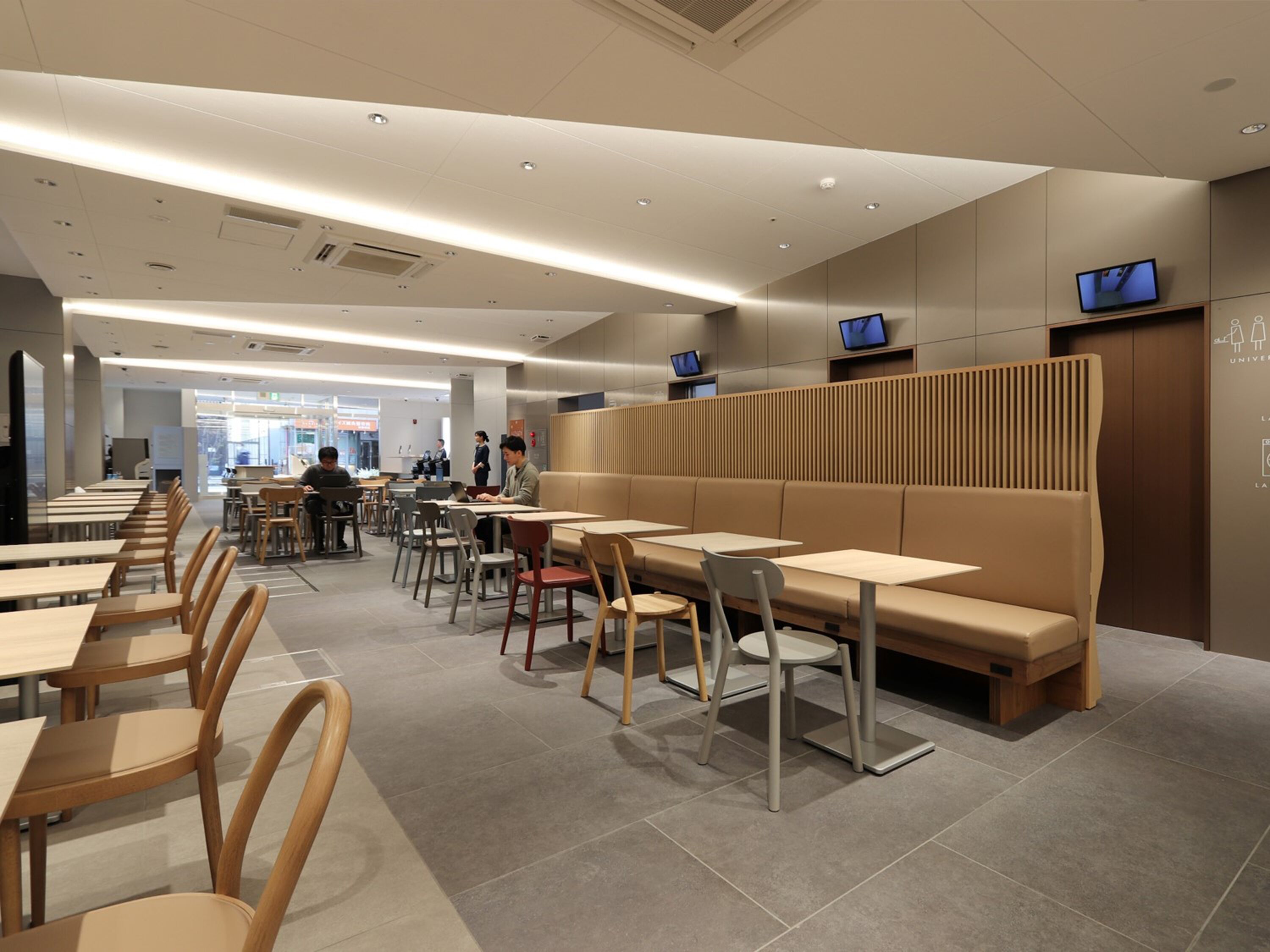 A modern dining area with long tables and benches, ceiling lights, and a few people in the background.