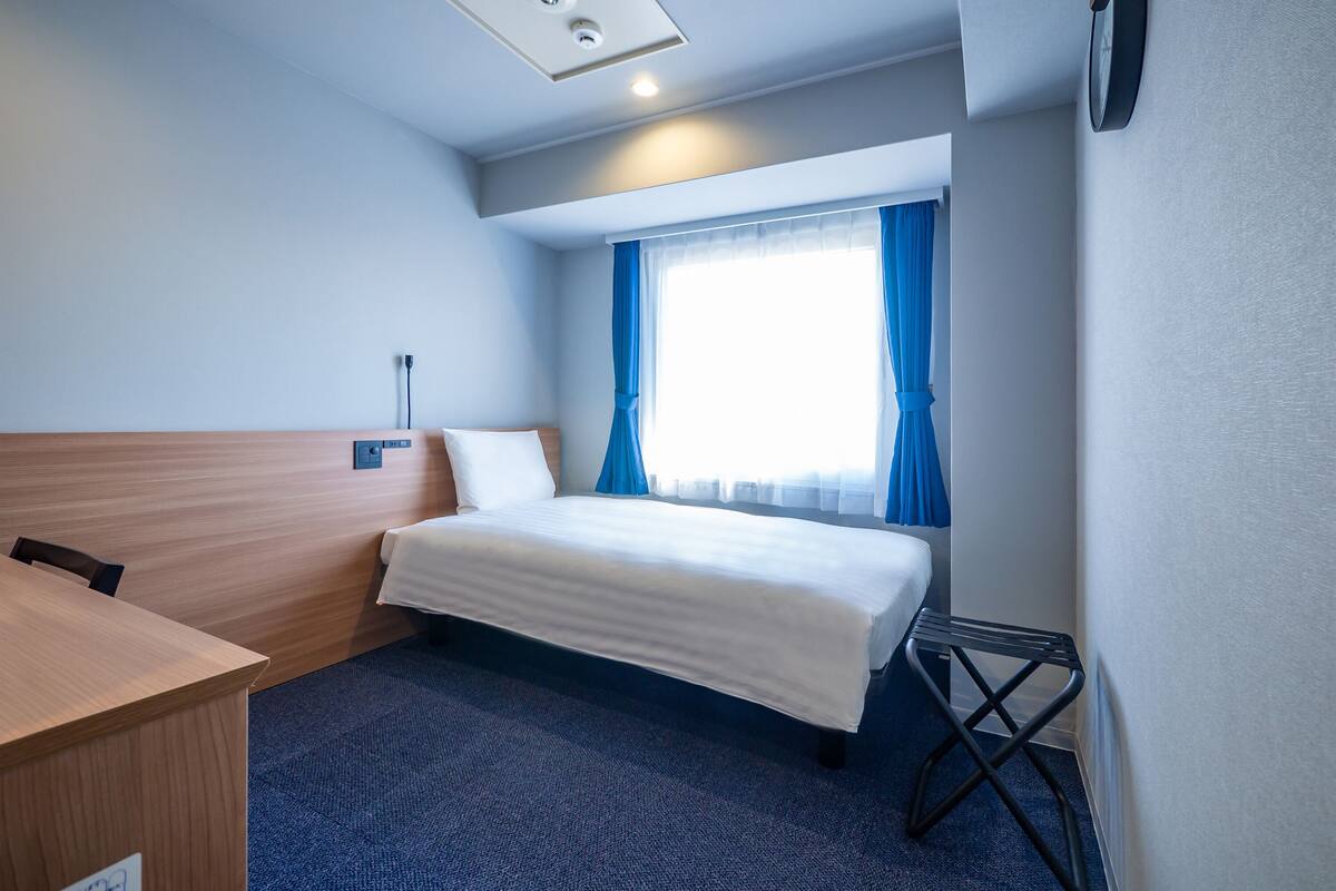 Una habitación de hotel con cama, escritorio, silla y ventana con cortinas.
