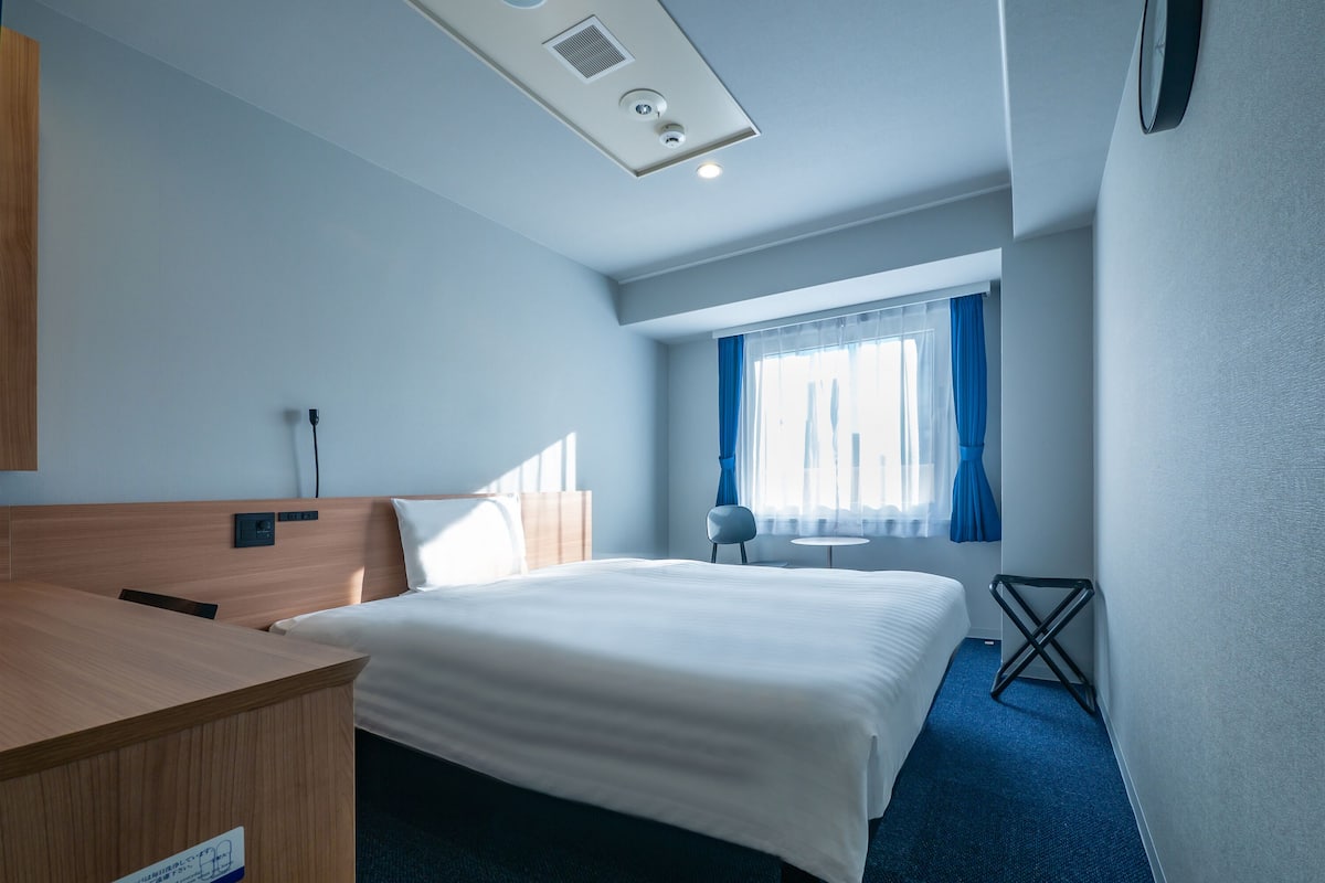 Habitación de hotel con cama, escritorio, silla, mesita, ventana con cortinas y un reloj en la pared.