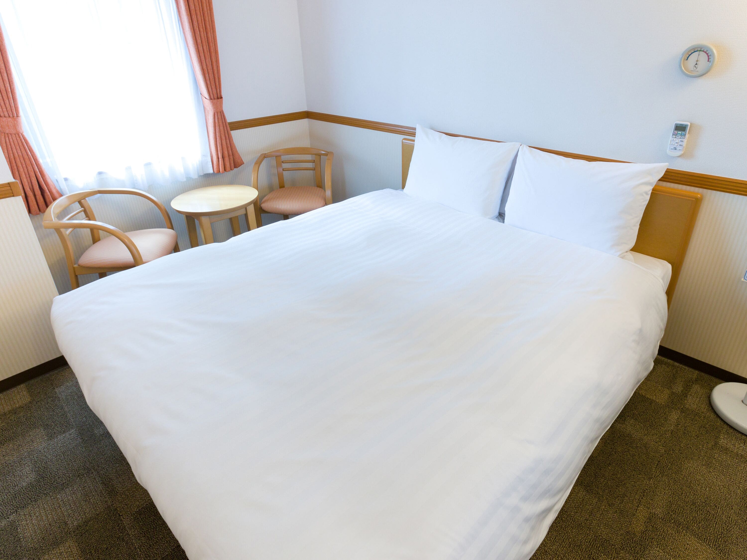 Een hotelkamer met een groot bed, twee stoelen, een klein tafeltje en een thermostaat aan de muur.