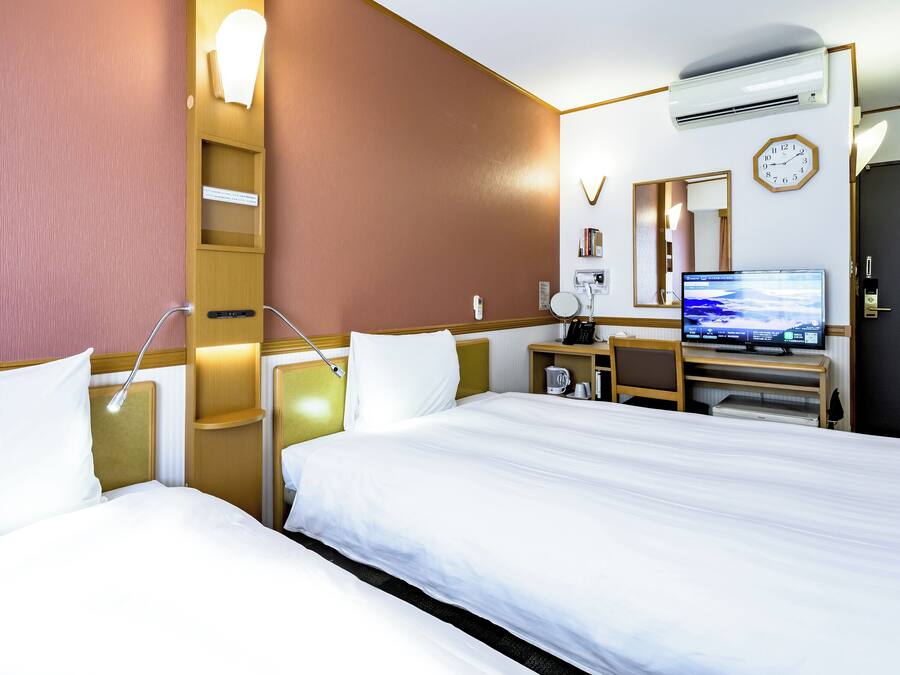 A hotel room with two beds, a desk with a computer, a clock, and an air conditioning unit.