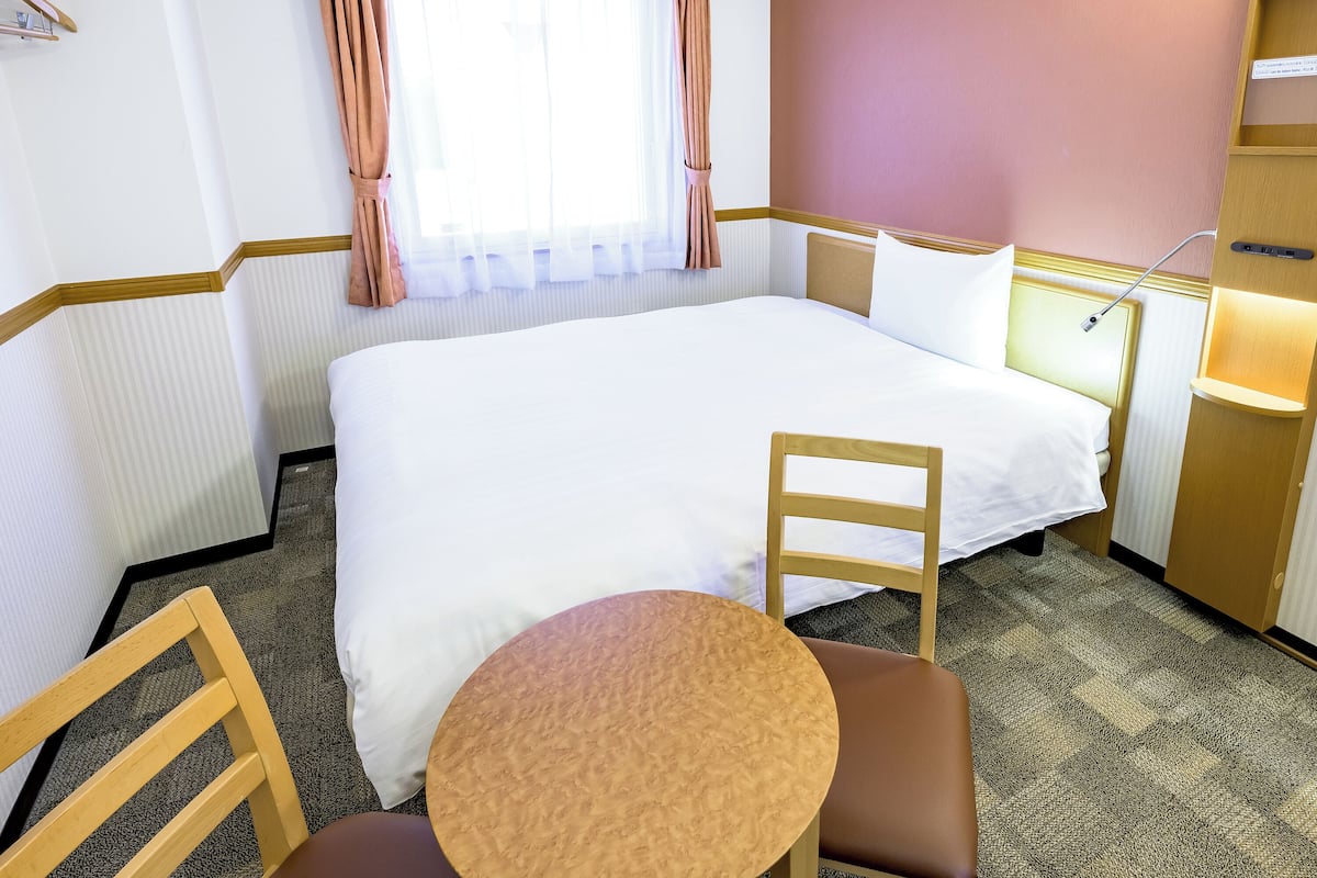 A hotel room with a large bed, a wooden chair, a small round table, and a nightstand with a lamp.