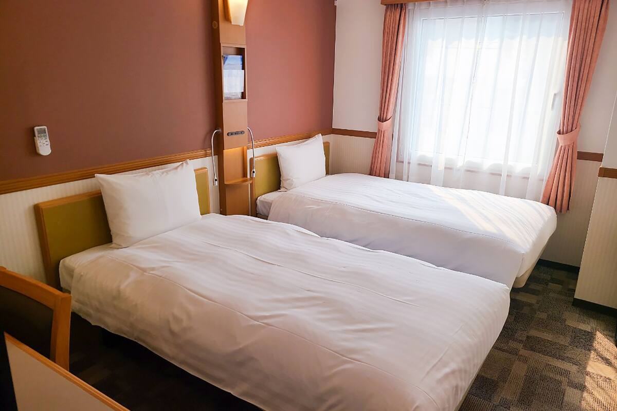 Une chambre d’hôtel avec deux lits, une table de chevet, une lampe, une fenêtre avec des rideaux et une télécommande fixée au mur.