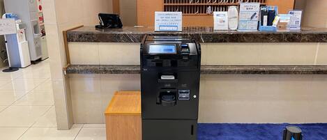 Zona de recepción de hotel con quiosco digital, caja registradora y señalización del punto de entrega de habitaciones.
