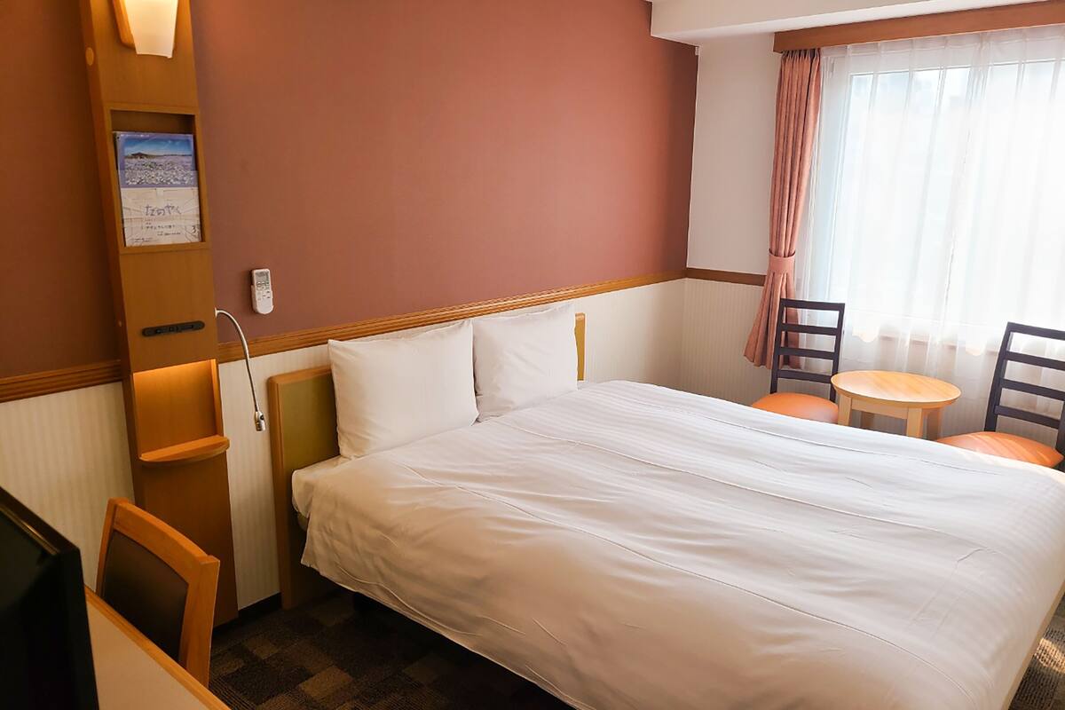 Une chambre d’hôtel avec un grand lit, un bureau et une chaise. Il y a une fenêtre avec des rideaux, un téléphone fixé au mur et une carte accrochée au mur.