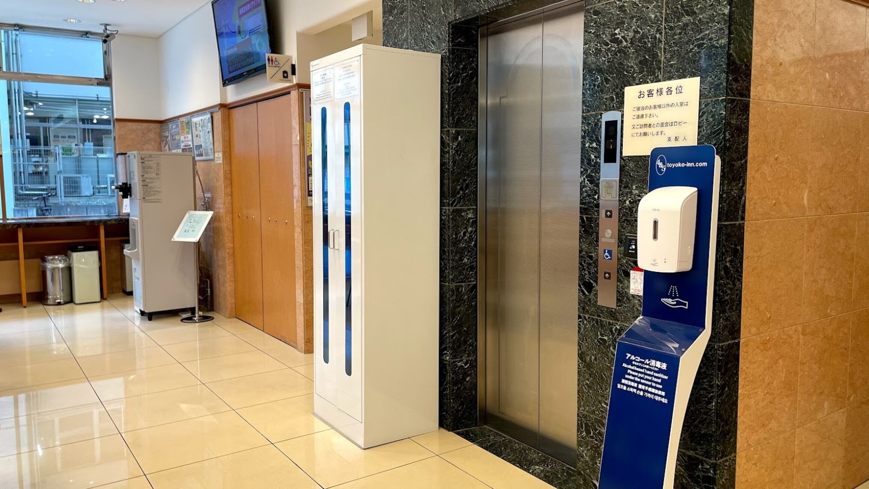 Um saguão de hotel com balcão de recepção, geladeira, máquina de venda automática e elevador.