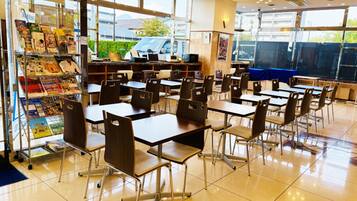 A spacious cafeteria with multiple tables and chairs, large windows, and a vending machine.