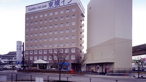 A multi-story hotel with a sign reading "toyoko-inn.com" and Japanese characters. It has a modern architectural style with a flat roof and a large vertical structure to its right. - Toyoko Inn Sakudaira Station Asama (Saku)