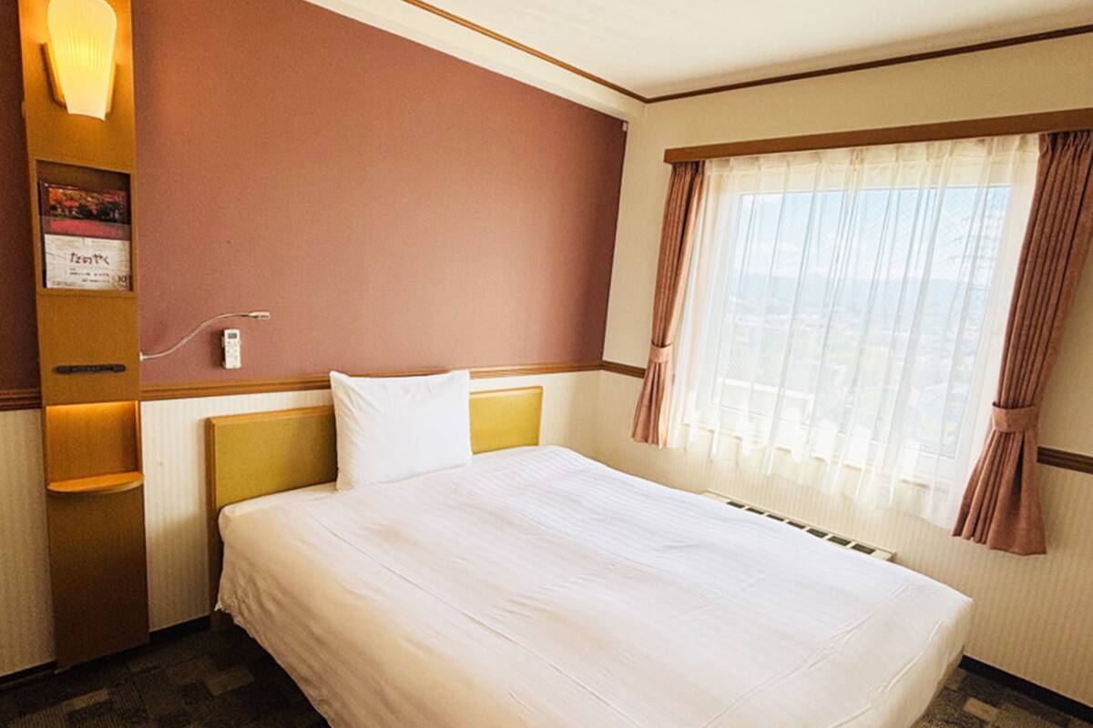 A hotel room with a large bed, a nightstand, a wall-mounted lamp, and a window with curtains.