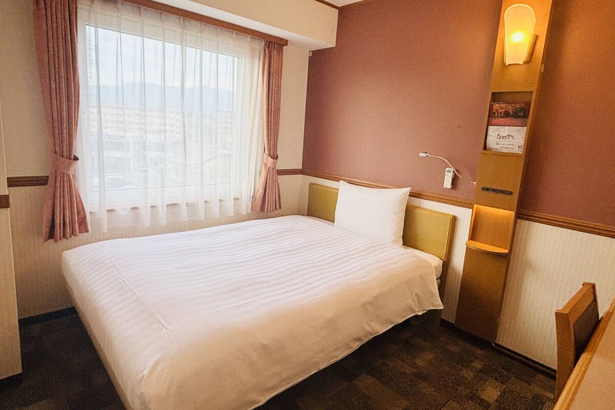 A hotel room with a large bed, a window with curtains, a wall-mounted light, and a small table with a lamp.