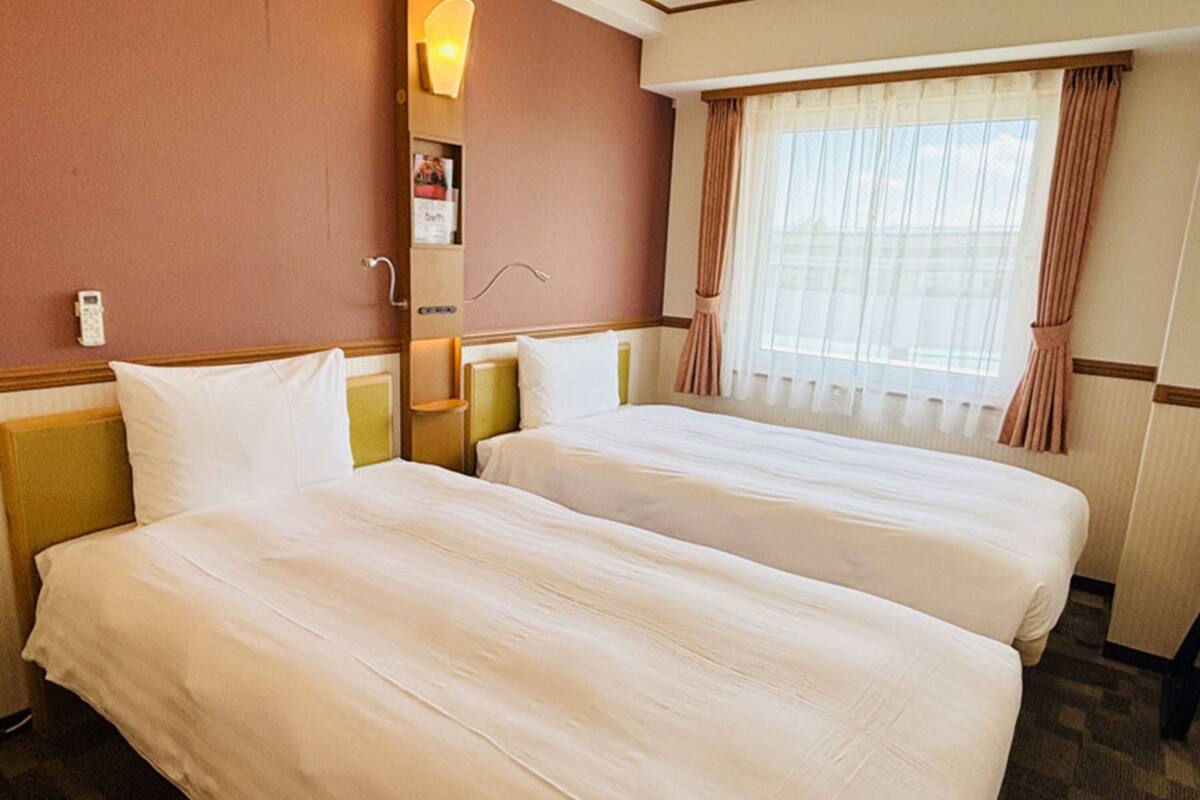 A hotel room with two beds, a bedside table, a lamp, a window with curtains, and a wall-mounted phone.