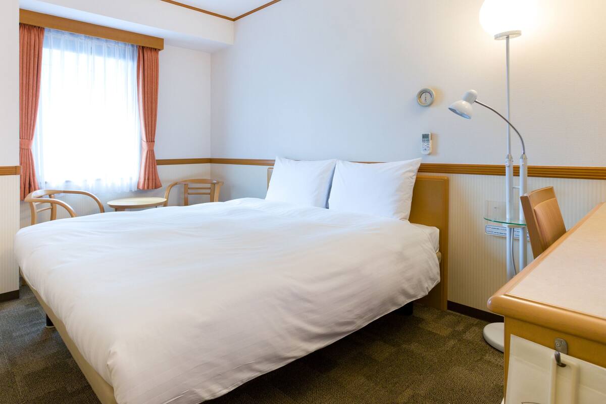 A hotel room with a large bed, a desk, a chair, a lamp, and a window with curtains.