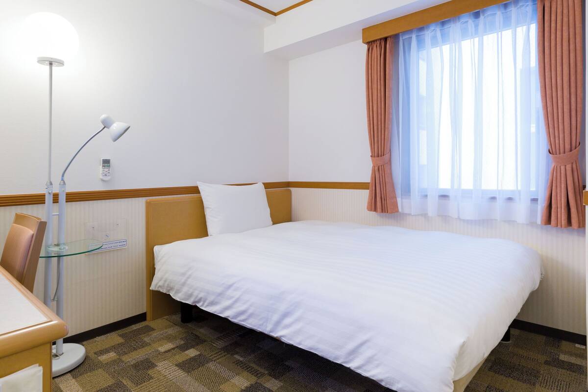 A hotel room with a bed, a desk, a lamp, and a window with curtains.