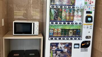 Máquina expendedora con diversas bebidas y aperitivos, un microondas y botes de basura.