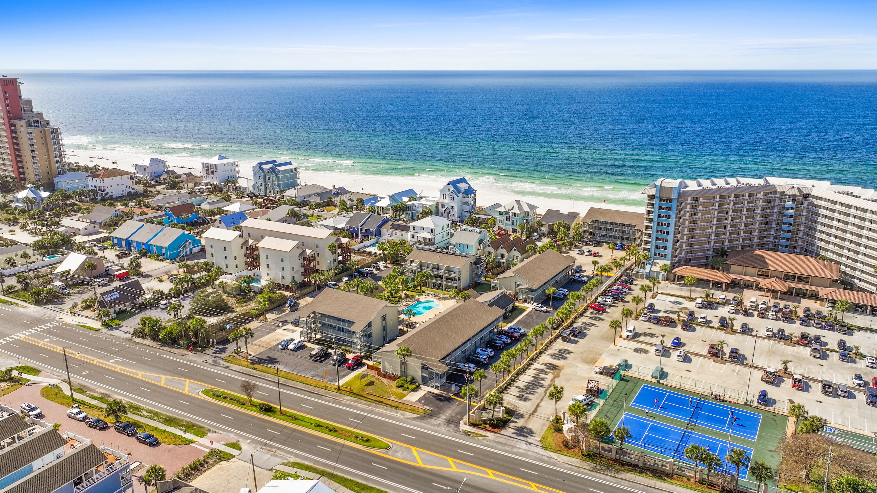 Vista aérea de un pueblo costero con hoteles frente a la playa, una cancha de tenis y una carretera con vehículos.