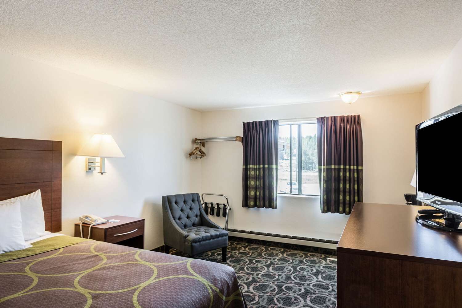 Quarto de hotel com cama, uma cadeira, escrivaninha com televisão, janela com cortinas e luminária na parede.