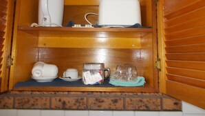 Un mueble de madera con dos estantes, que contiene una tetera eléctrica blanca, una cafetera blanca, un tarro de vidrio con una bolsita de té, una taza de vidrio y un paño verde doblado.