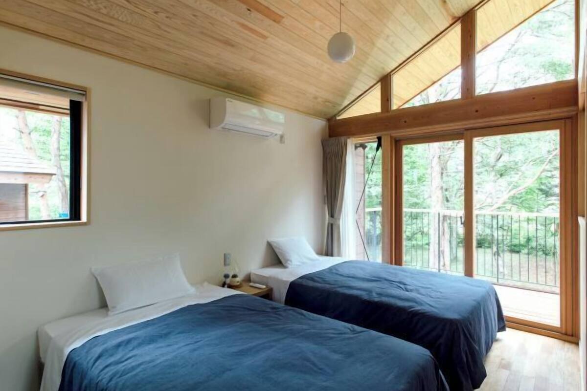A bedroom with two beds, a wooden ceiling, large windows, and an air conditioning unit.