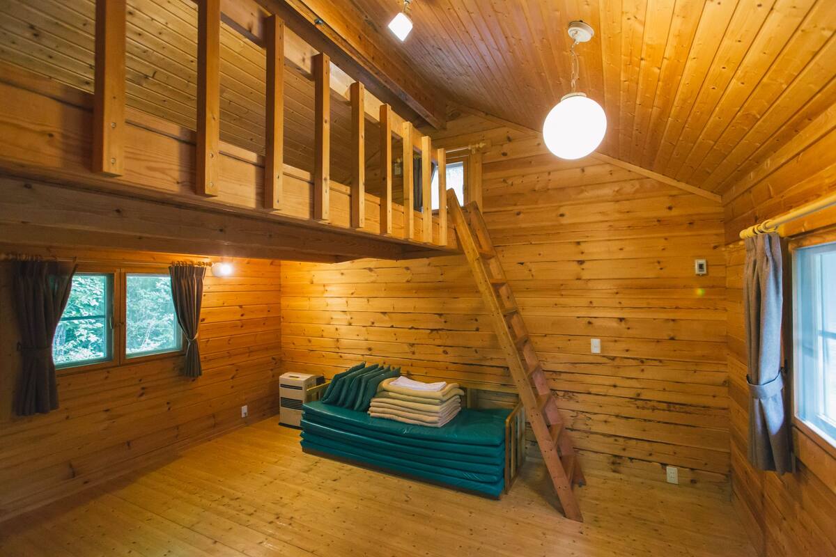 A wooden cabin interior with a loft area, a staircase, and a window with curtains.